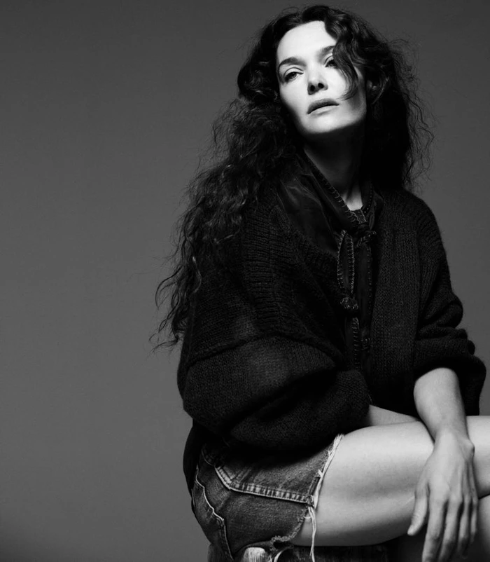 Monochrome portrait of Ana Rujas with long curly dark hair wearing a dark knitted cardigan and denim shorts against a grey studio background