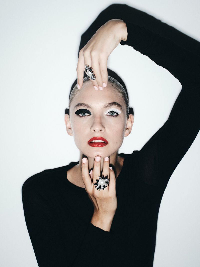 Model in black top wearing statement rings with dramatic black and white eye makeup against a white background