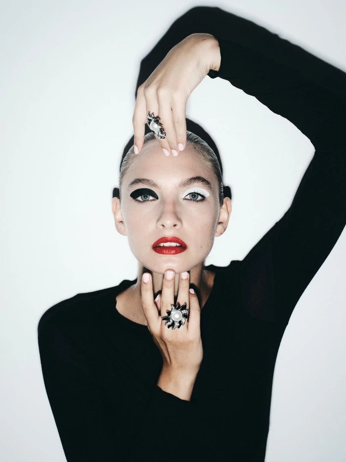 Model in black top wearing statement rings with dramatic black and white eye makeup against a white background