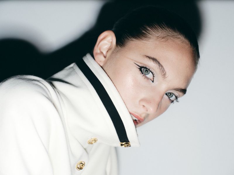 Close-up portrait of a model wearing a high-collared white coat with dark contrast trim against a bright wall.