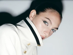 Close-up portrait of a model wearing a high-collared white coat with dark contrast trim against a bright wall.