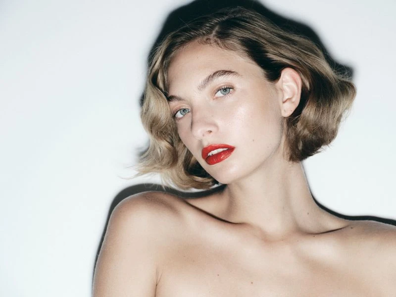 Close-up portrait of a woman with short blonde hair and bright red lipstick casting a sharp dark shadow against a white studio wall.