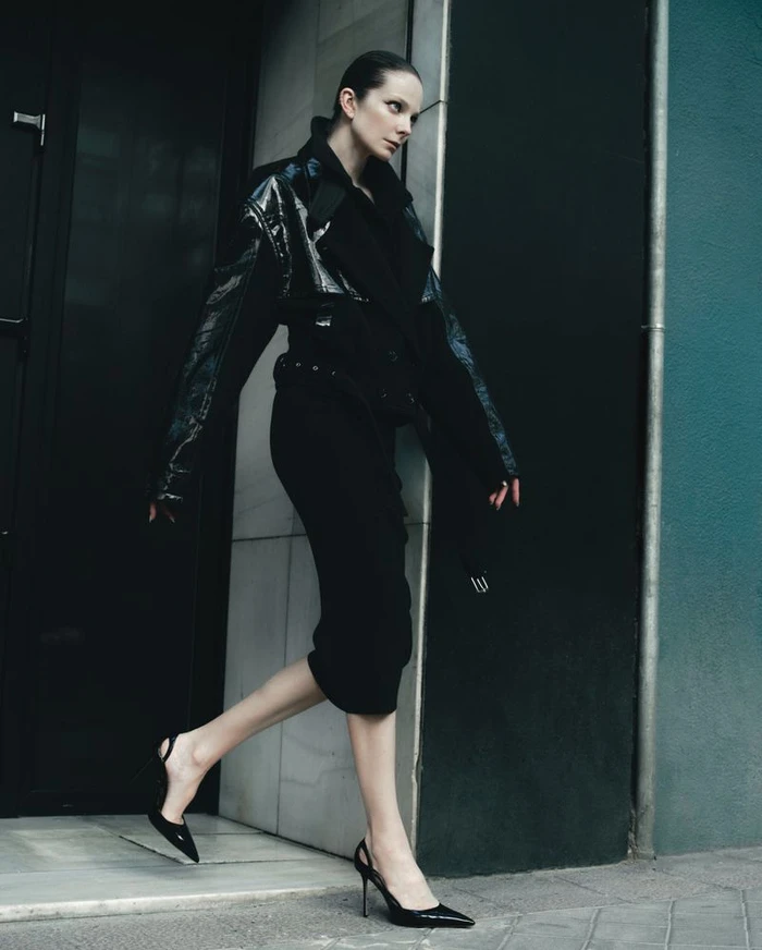 Model in black jacket and skirt walking past a dark architectural building exterior