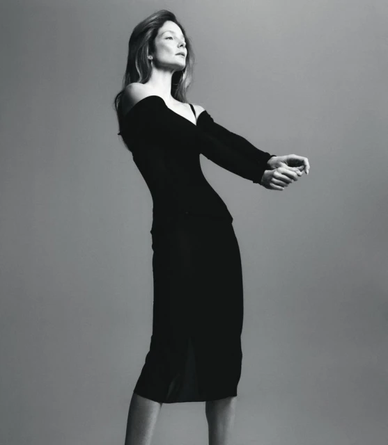 Black and white editorial portrait of model Eniko Mihalik wearing an off-the-shoulder black dress against a plain studio background.