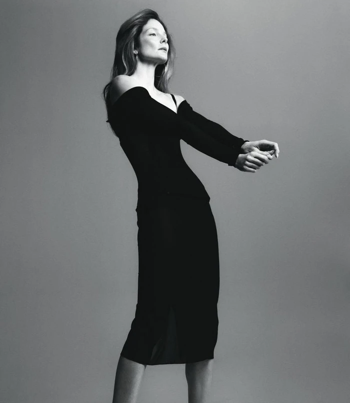 Black and white editorial portrait of model Eniko Mihalik wearing an off-the-shoulder black dress against a plain studio background.