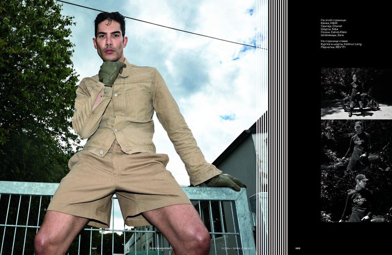 Man wearing a khaki jacket and shorts with gloves outdoors leaning on a metal fence for POSE Magazine