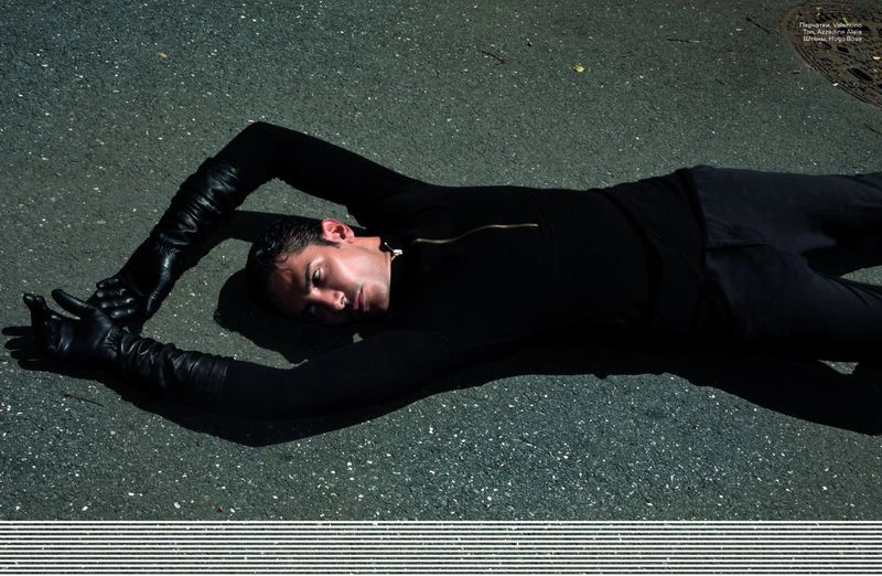 Man lying on asphalt ground wearing a black turtleneck and long black leather gloves with arms raised above his head