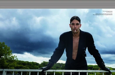 Man in a dark suit jacket and leather gloves leaning on a railing against a dramatic cloudy sky.