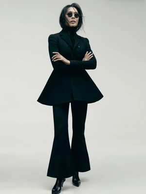 Model with dark hair and sunglasses wearing a black peplum jacket and trousers against a light backdrop