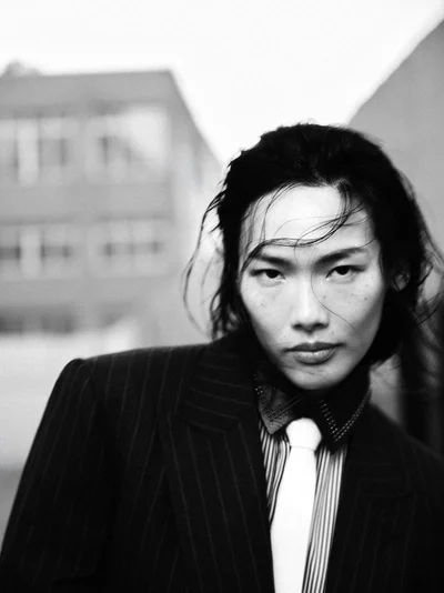 Monochrome portrait of a model in a pinstripe blazer, collared shirt, and white tie against an out-of-focus urban background.