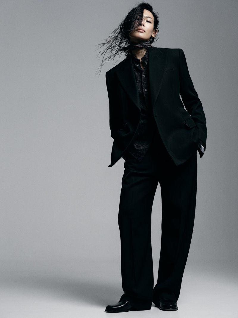 Model in black suit standing against a grey studio backdrop with dark hair blowing across her face