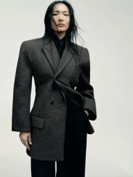 Model with dark flowing hair wearing a structured grey blazer and black trousers against a neutral background.
