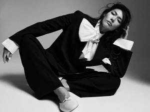 Model wearing a black suit and white shirt with a large bow sitting on a grey studio seamless background.