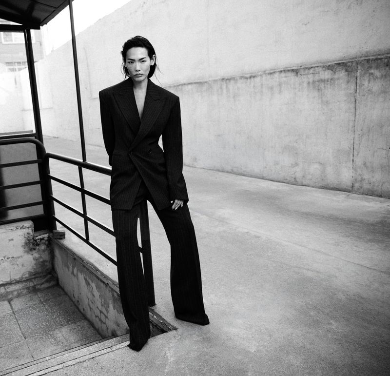 Model standing outdoors wearing a dark structured blazer and matching trousers leaning against a railing