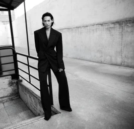 Model standing outdoors wearing a dark structured blazer and matching trousers leaning against a railing