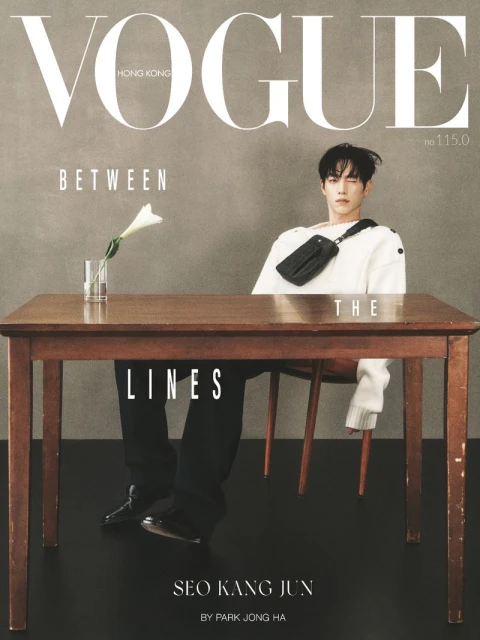 Man seated behind a large wooden table with a flower in a glass, wearing a white sweater and black crossbody bag against a grey textured wall.