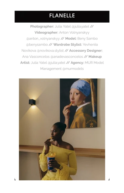 Black woman in a yellow off-shoulder top holding a blue cup against a wall with a painting and modern wall sconce.
