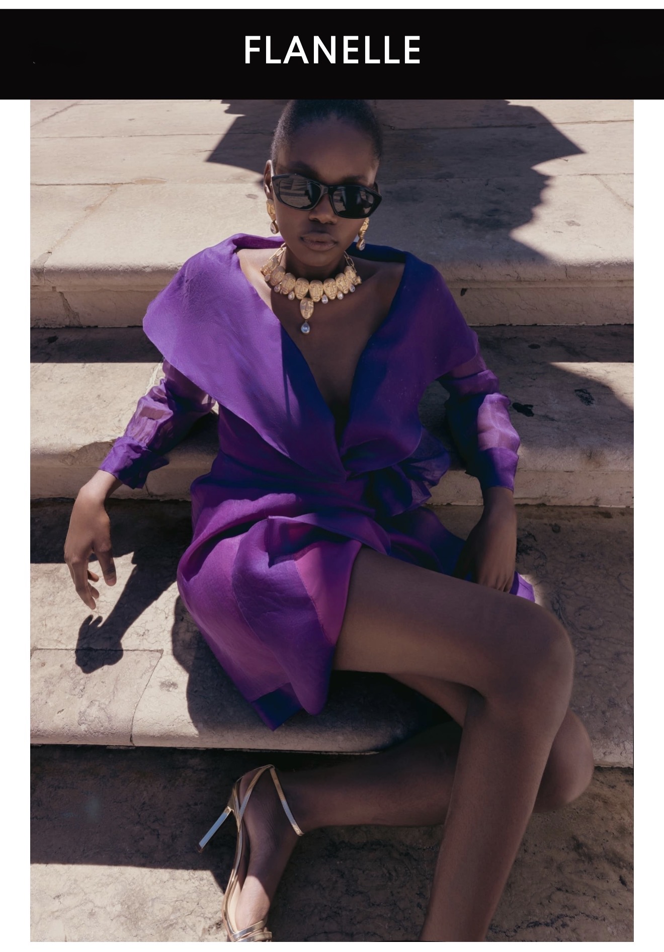 Model wearing a purple dress and sunglasses seated on stone steps outdoors under natural sunlight