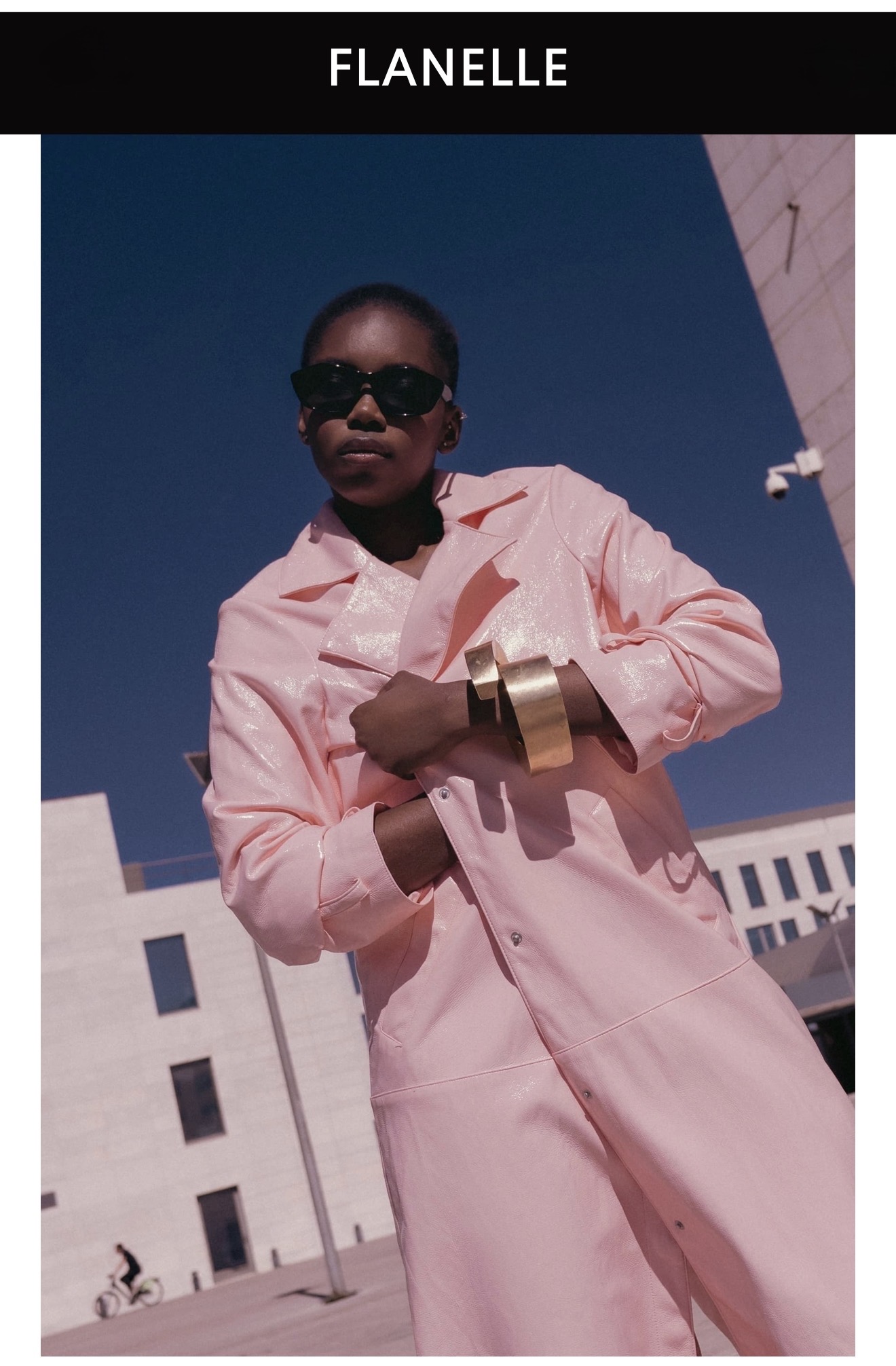 Model in dark sunglasses wearing a pink textured coat and gold cuffs outdoors against a modern building exterior.