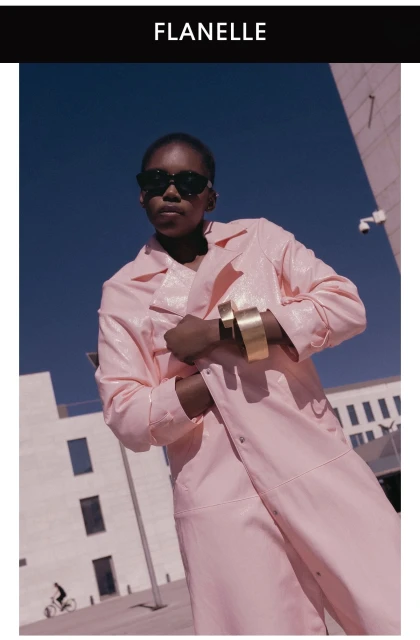 Model in dark sunglasses wearing a pink textured coat and gold cuffs outdoors against a modern building exterior.