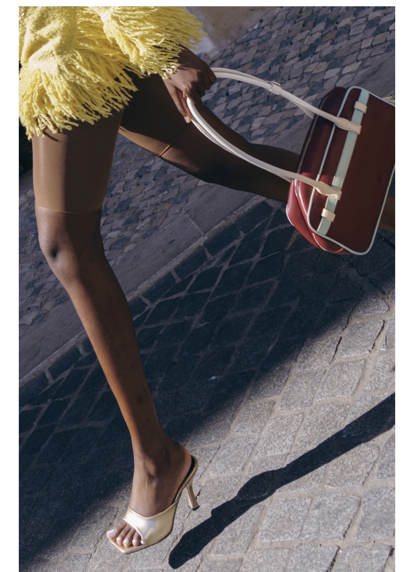 Model wearing a bright yellow fringed top and metallic heeled sandals while walking on cobblestone street carrying a handbag.