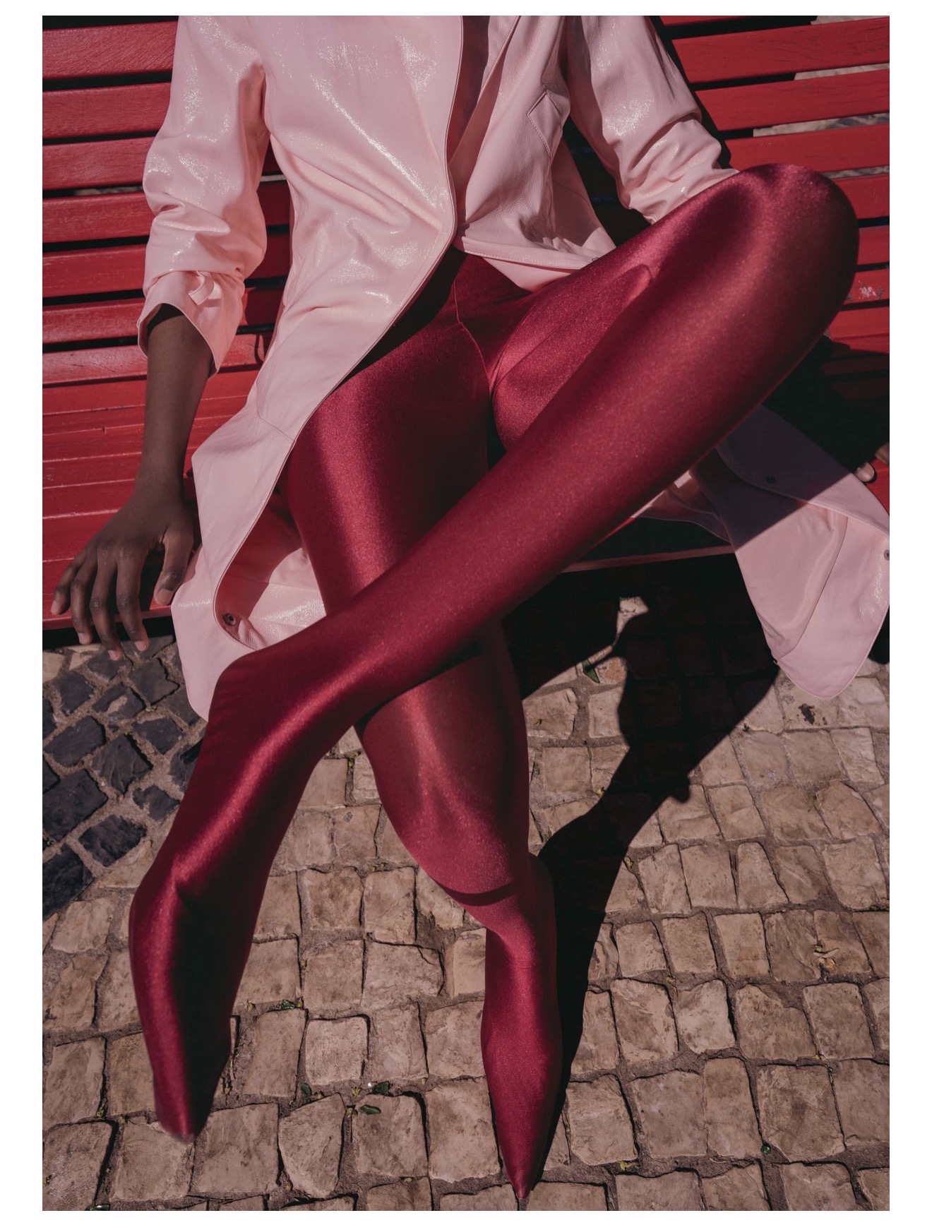Model wearing shiny red leggings and a light pink coat seated on a red bench over cobblestone