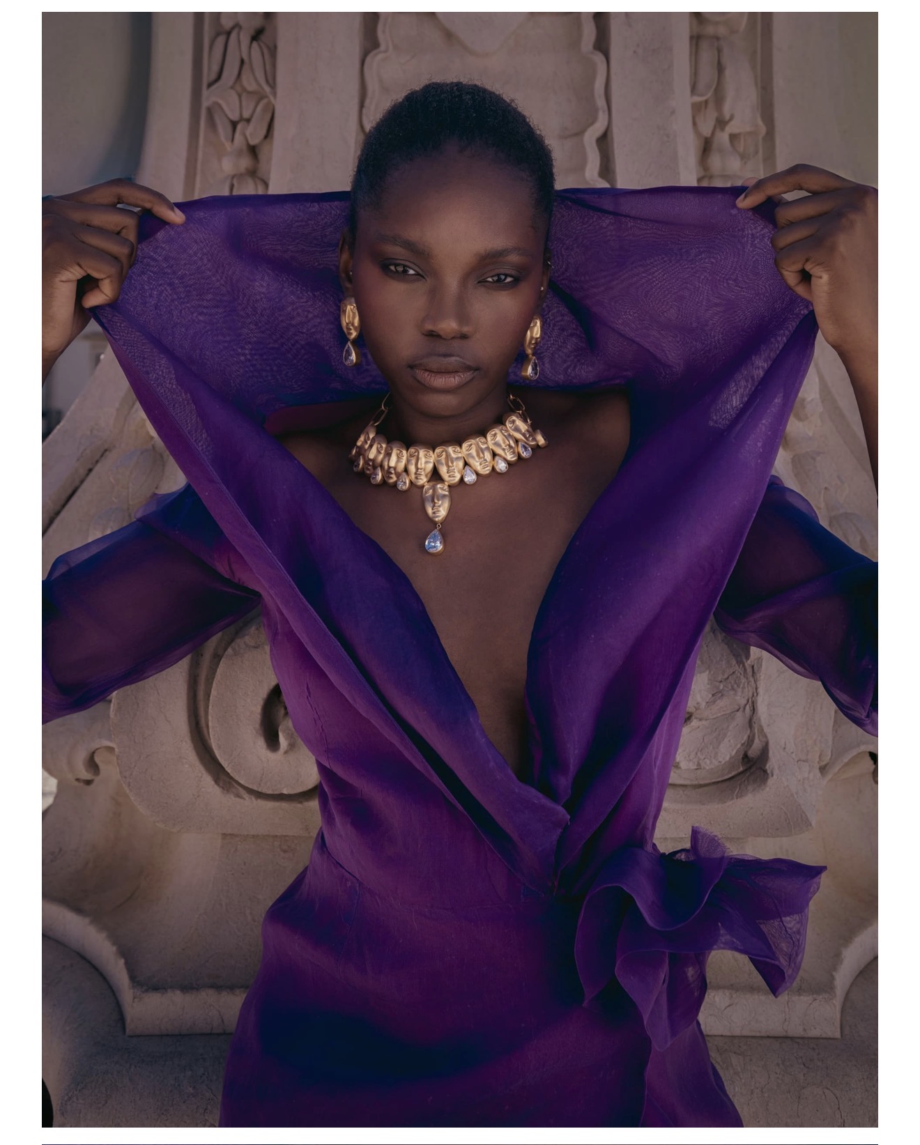Black woman wearing a deep purple dress and gold statement necklace holding a matching purple fabric overhead