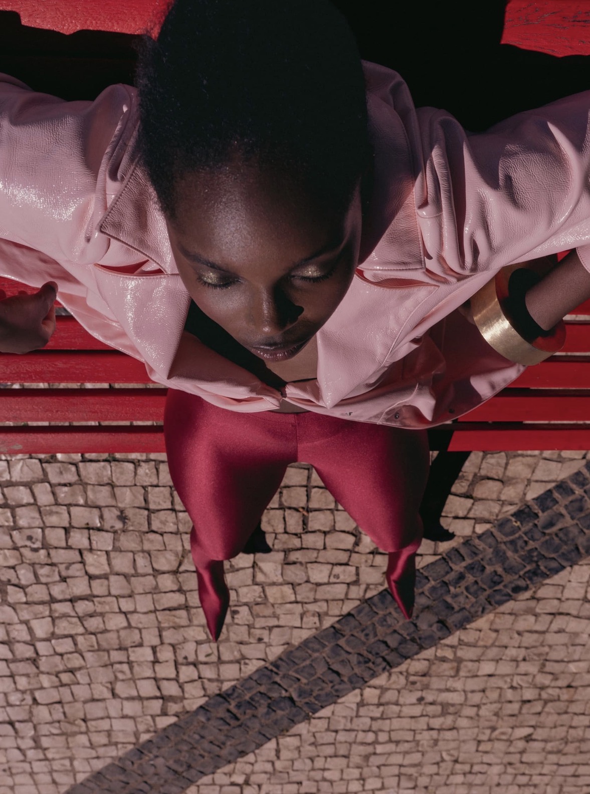 High-angle shot of a black woman wearing a pink jacket and burgundy trousers, seated on a red bench.