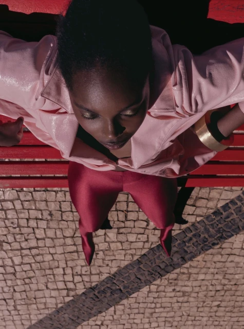 High-angle shot of a black woman wearing a pink jacket and burgundy trousers, seated on a red bench.