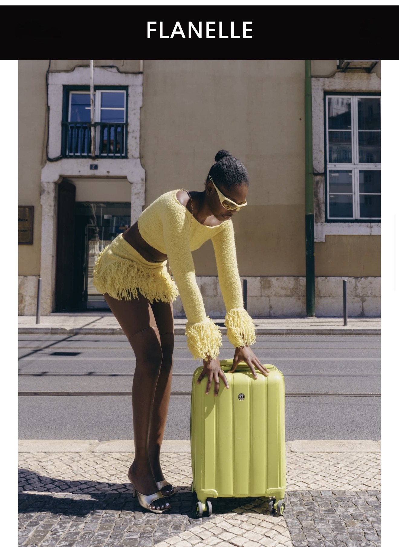 Black woman wearing a yellow textured two-piece outfit and sunglasses leaning on a matching green suitcase on a street.
