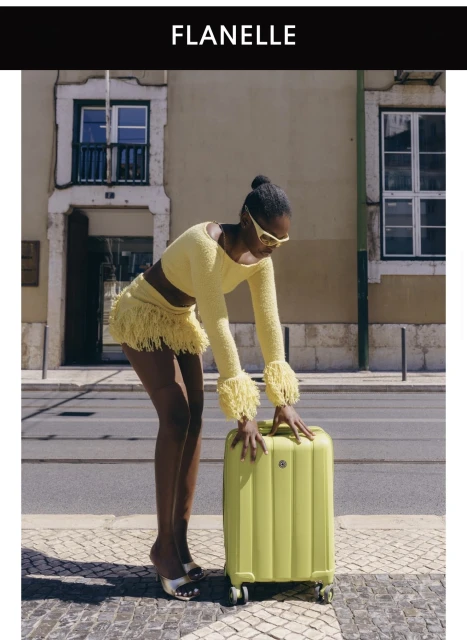 Black woman wearing a yellow textured two-piece outfit and sunglasses leaning on a matching green suitcase on a street.