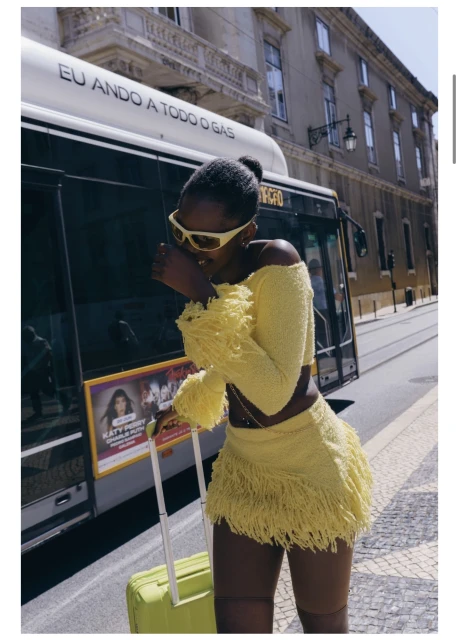 Black woman in a yellow fringed two-piece outfit holding a matching suitcase in front of a bus