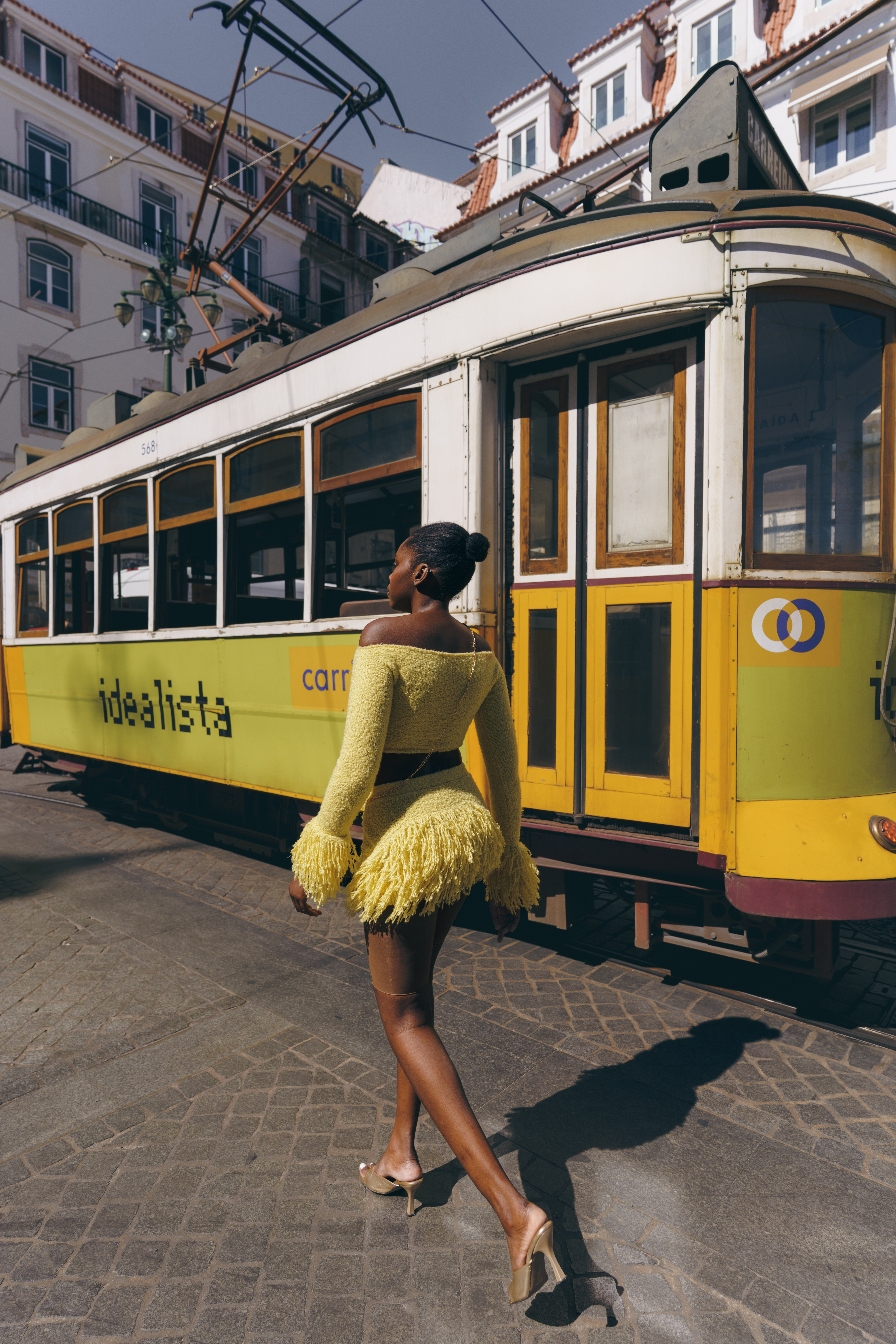 Black woman walking past a yellow vintage tram wearing a yellow textured off-shoulder dress in Lisbon.