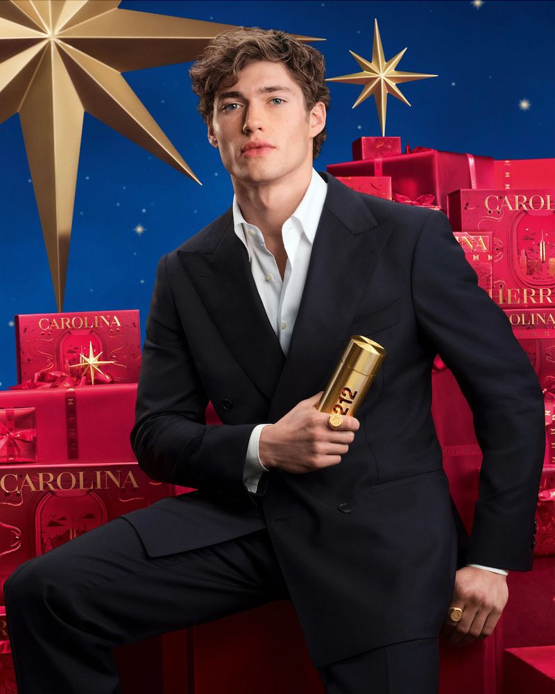 Josh Upshaw in a black suit holding a gold 212 perfume bottle against a holiday backdrop