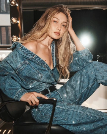 Gigi Hadid wearing a denim embellished jumpsuit seated in a chair by a lit makeup mirror