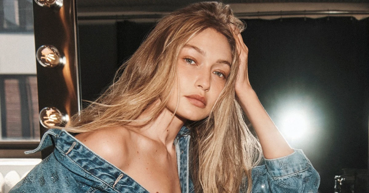 Gigi Hadid in a denim jacket running her hand through her blonde hair beside an illuminated vanity mirror.