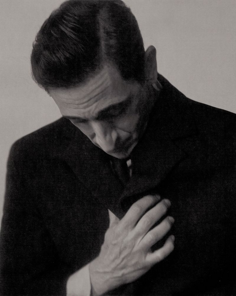Close-up portrait of actor Adrien Brody in a dark coat looking downward with his hand on his chest against a neutral studio background