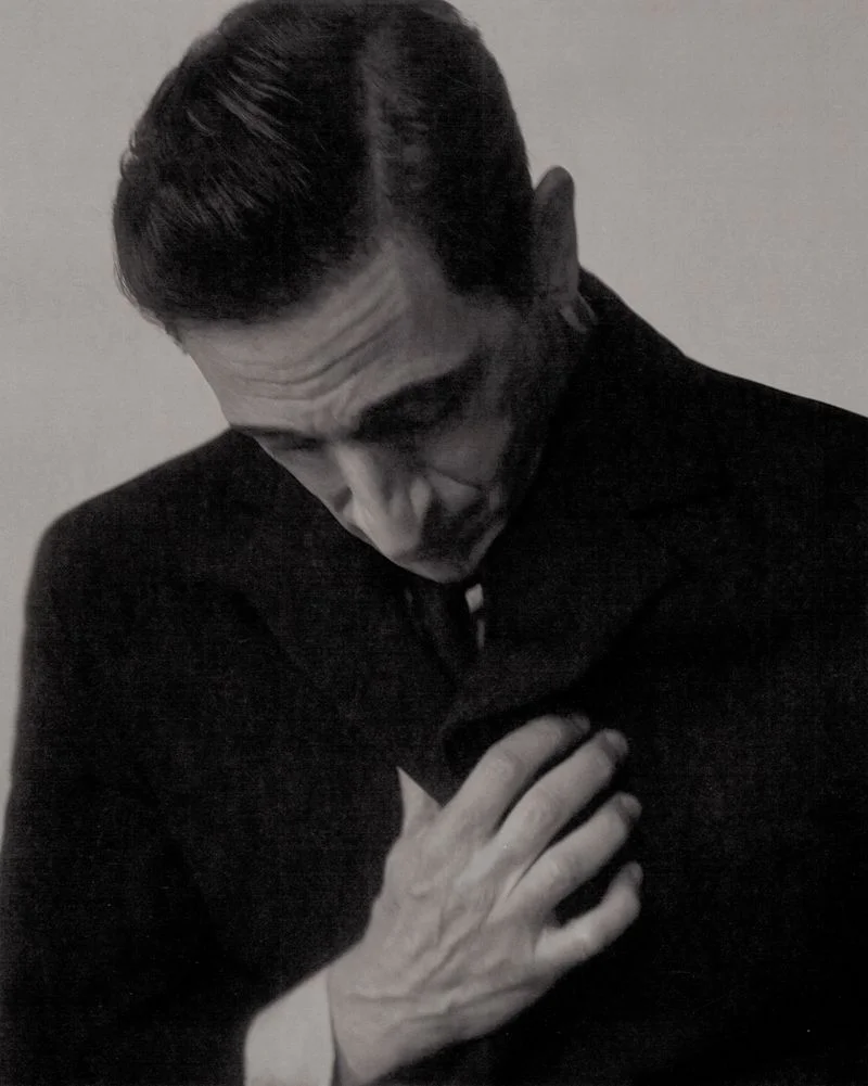 Close-up portrait of actor Adrien Brody in a dark coat looking downward with his hand on his chest against a neutral studio background