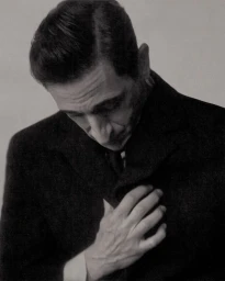 Close-up portrait of actor Adrien Brody in a dark coat looking downward with his hand on his chest against a neutral studio background