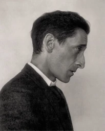 Profile portrait of Adrien Brody wearing a dark jacket and white shirt against a neutral background