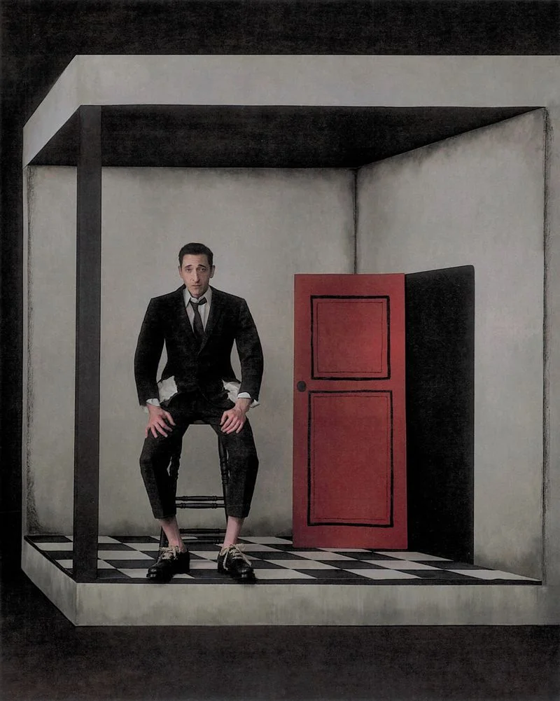 Actor Adrien Brody seated on a stool inside a surreal architectural room with a black and white tiled floor and a red door.