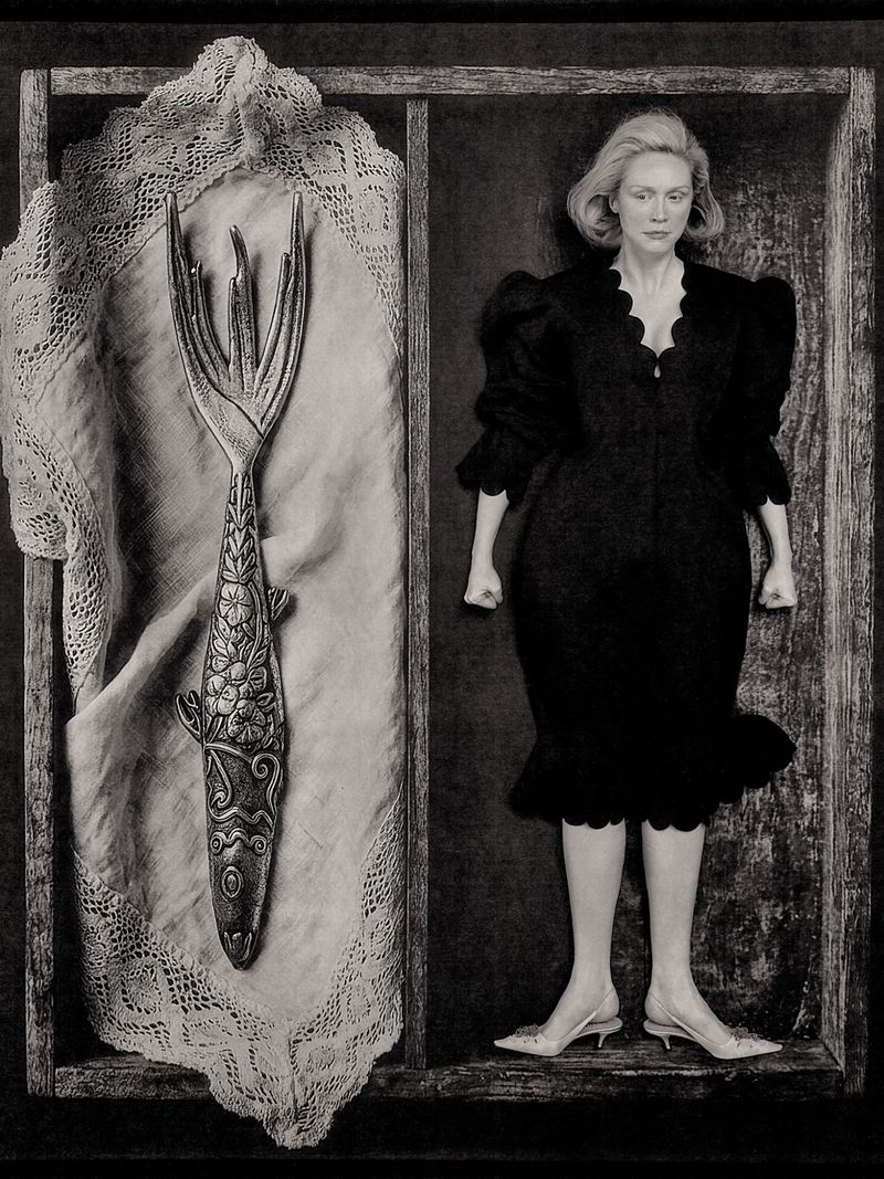 Gwendoline Christie wearing a black dress inside a wooden compartment beside a large decorative fish fork resting on lace fabric