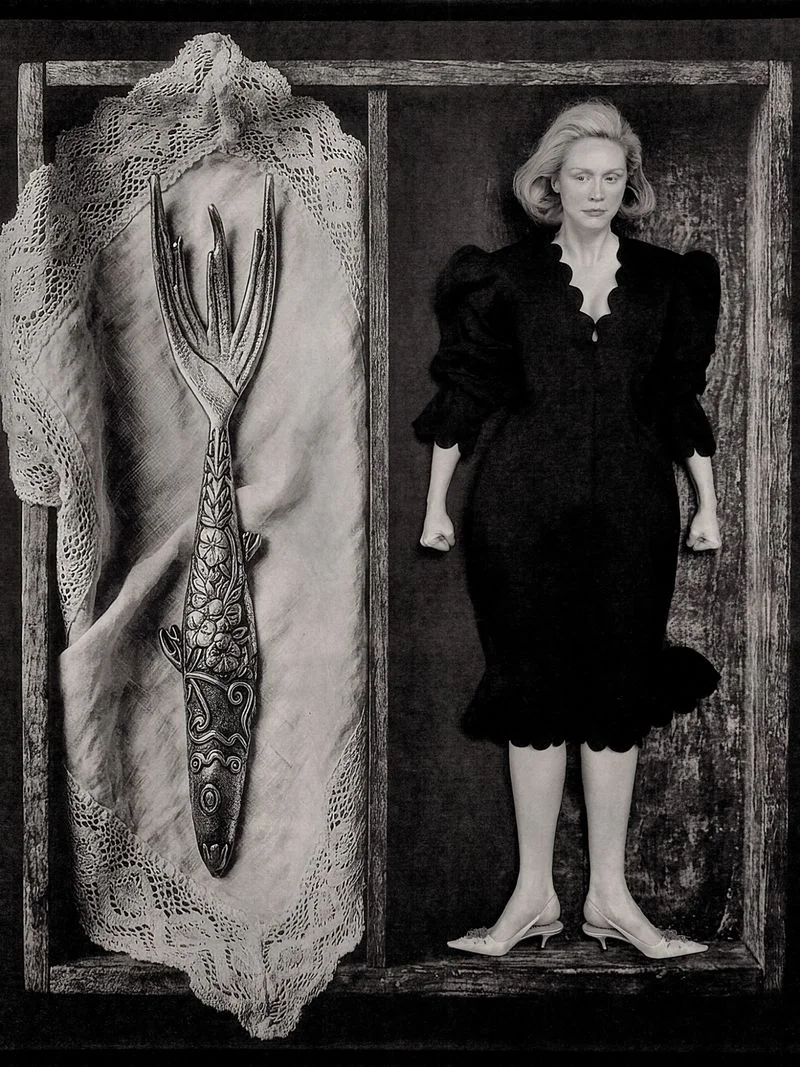 Gwendoline Christie wearing a black dress inside a wooden compartment beside a large decorative fish fork resting on lace fabric