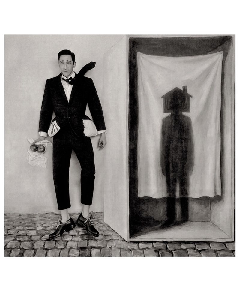 Adrien Brody in a dark suit standing next to a framed structure containing a curtained shadow silhouette