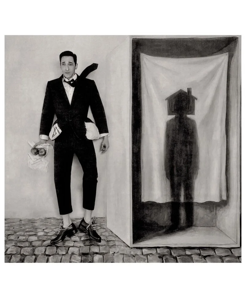 Adrien Brody in a dark suit standing next to a framed structure containing a curtained shadow silhouette
