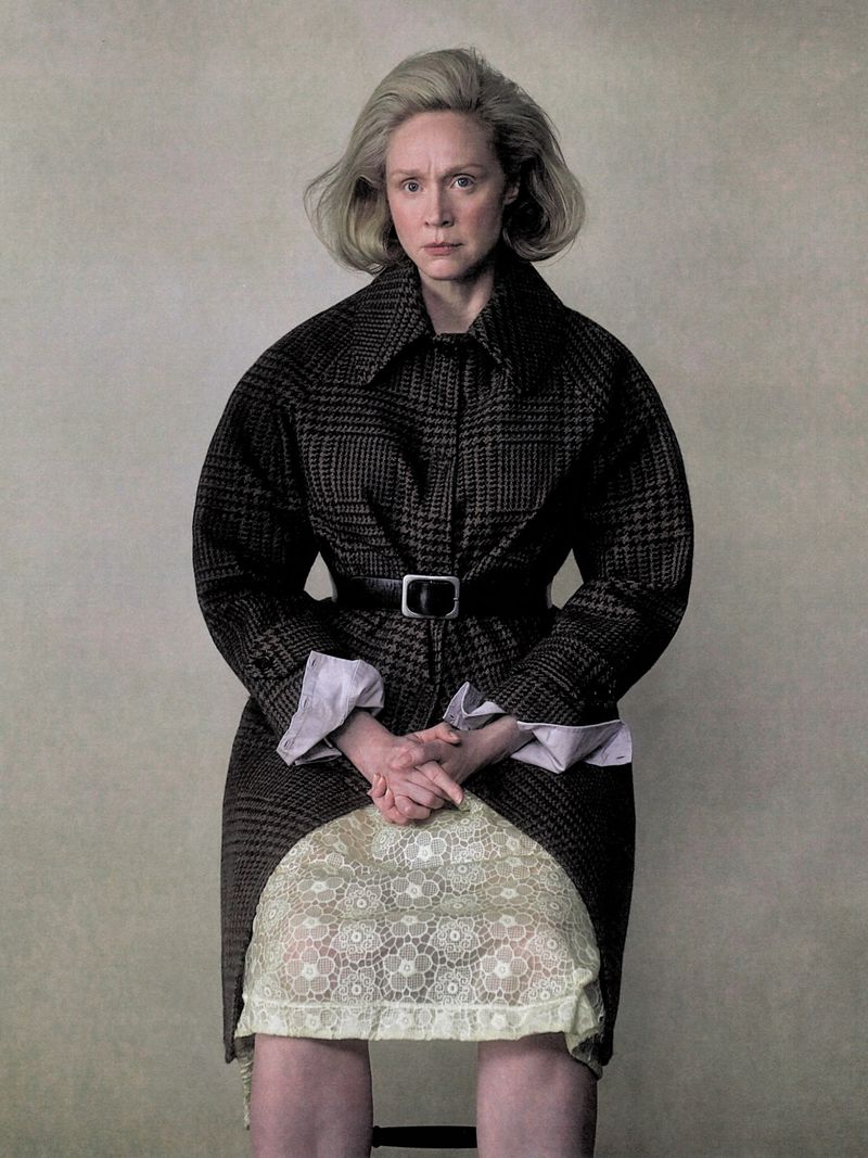Gwendoline Christie seated wearing a patterned belted coat over a textured lace skirt against a plain backdrop