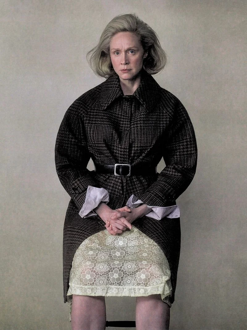 Gwendoline Christie seated wearing a patterned belted coat over a textured lace skirt against a plain backdrop