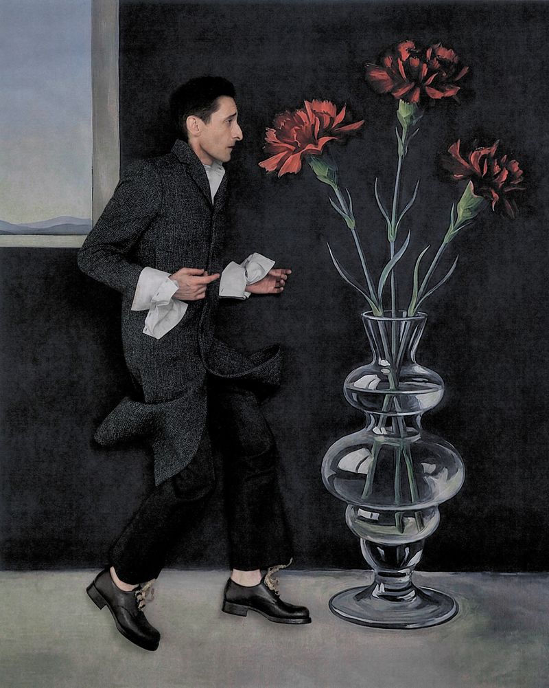 Adrien Brody in a grey coat beside a large glass vase with red flowers for Bauhaus Ballet