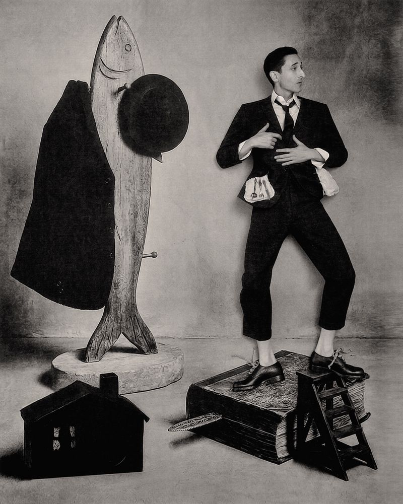 Adrien Brody posing beside a large wooden fish sculpture holding a hat in a monochrome editorial portrait.