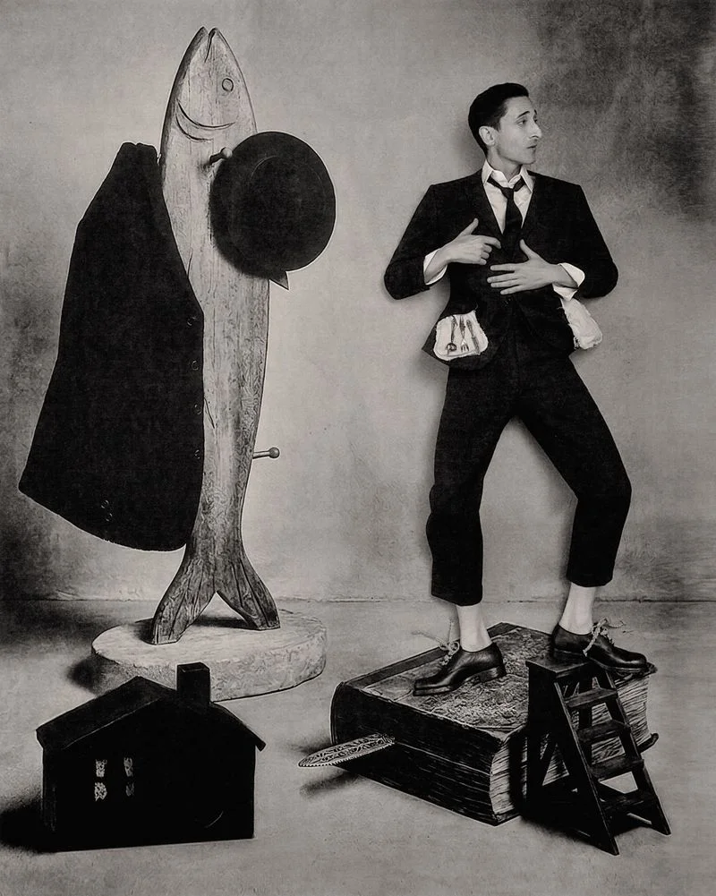 Adrien Brody posing beside a large wooden fish sculpture holding a hat in a monochrome editorial portrait.
